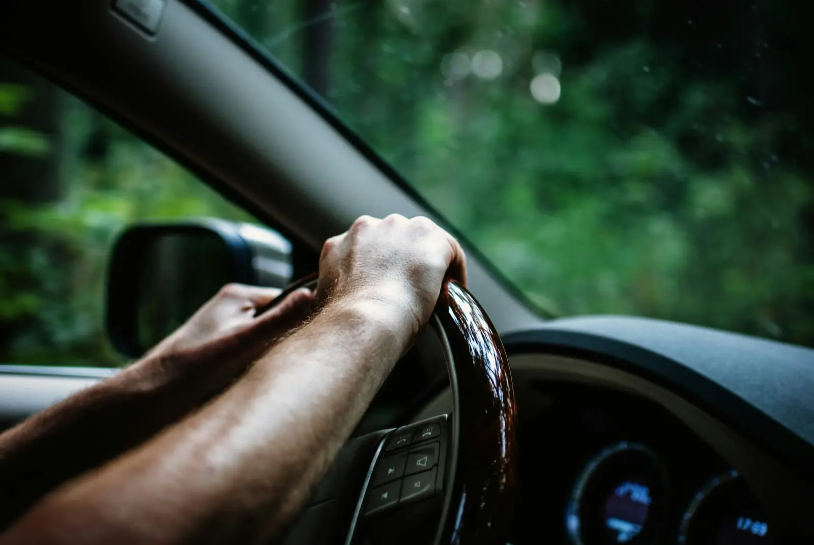 Hand eines Fahrers am Lenkrad, draußen Bäume im Tageslicht